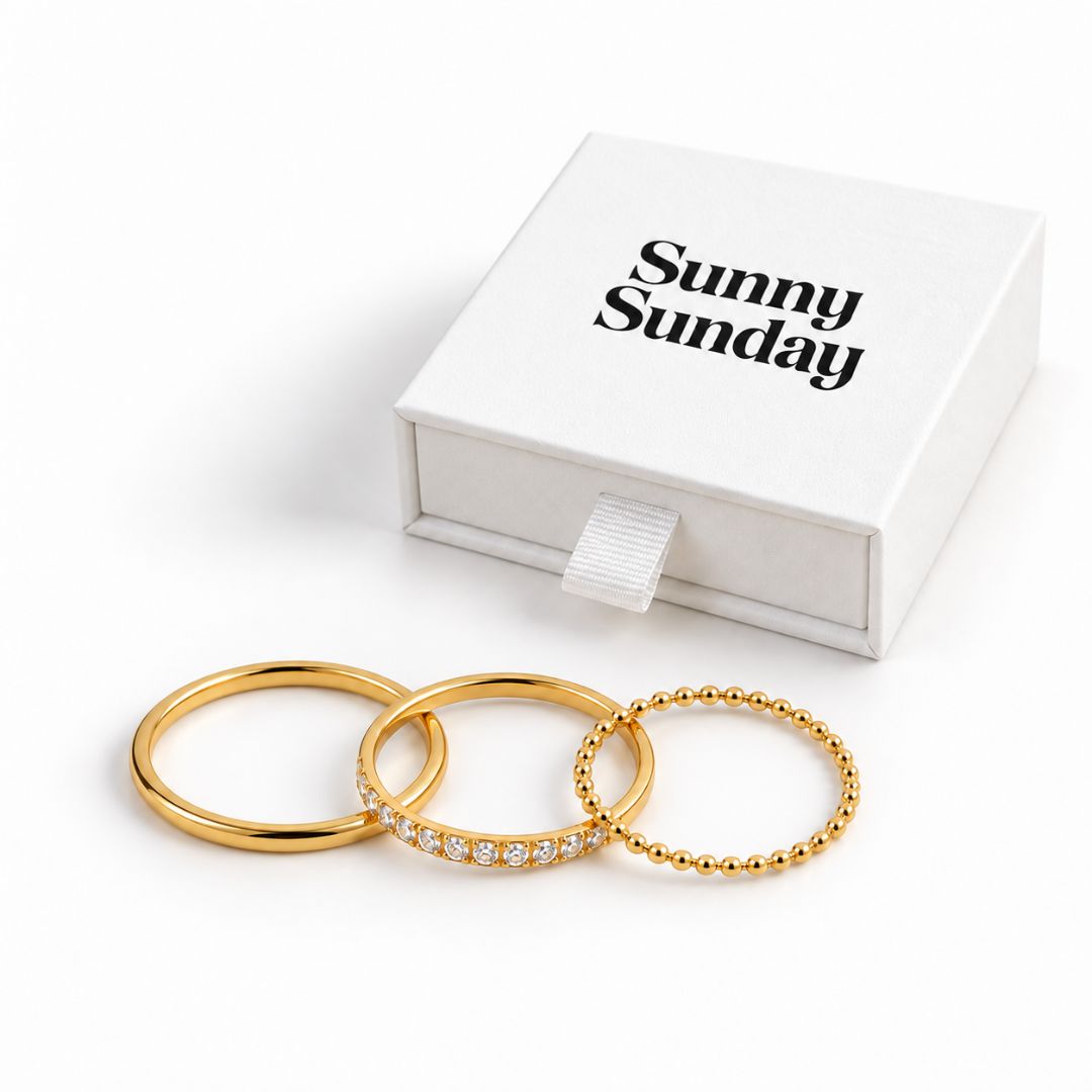 Three gold rings displayed in front of a white gift box with the text 'Sunny Sunday' on a clean white background. One ring is smooth, one is lined with small clear stones, and one has a beaded texture.