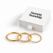 Three gold rings displayed in front of a white gift box with the text 'Sunny Sunday' on a clean white background. One ring is smooth, one is lined with small clear stones, and one has a beaded texture.