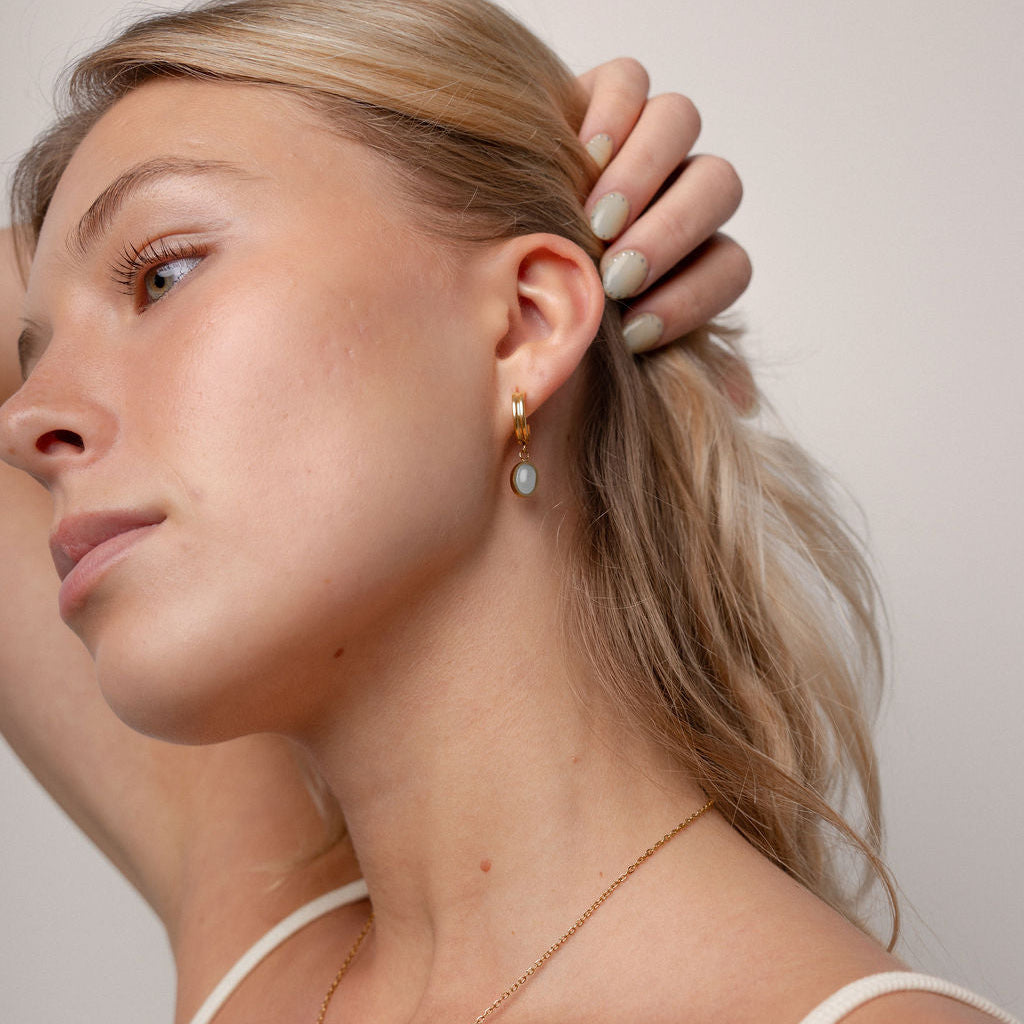 A close-up lifestyle shot of a blonde woman wearing a delicate gold necklace with a small green and gold pendant and matching drop earrings, paired with a cream-colored spaghetti strap top against a soft beige background.
