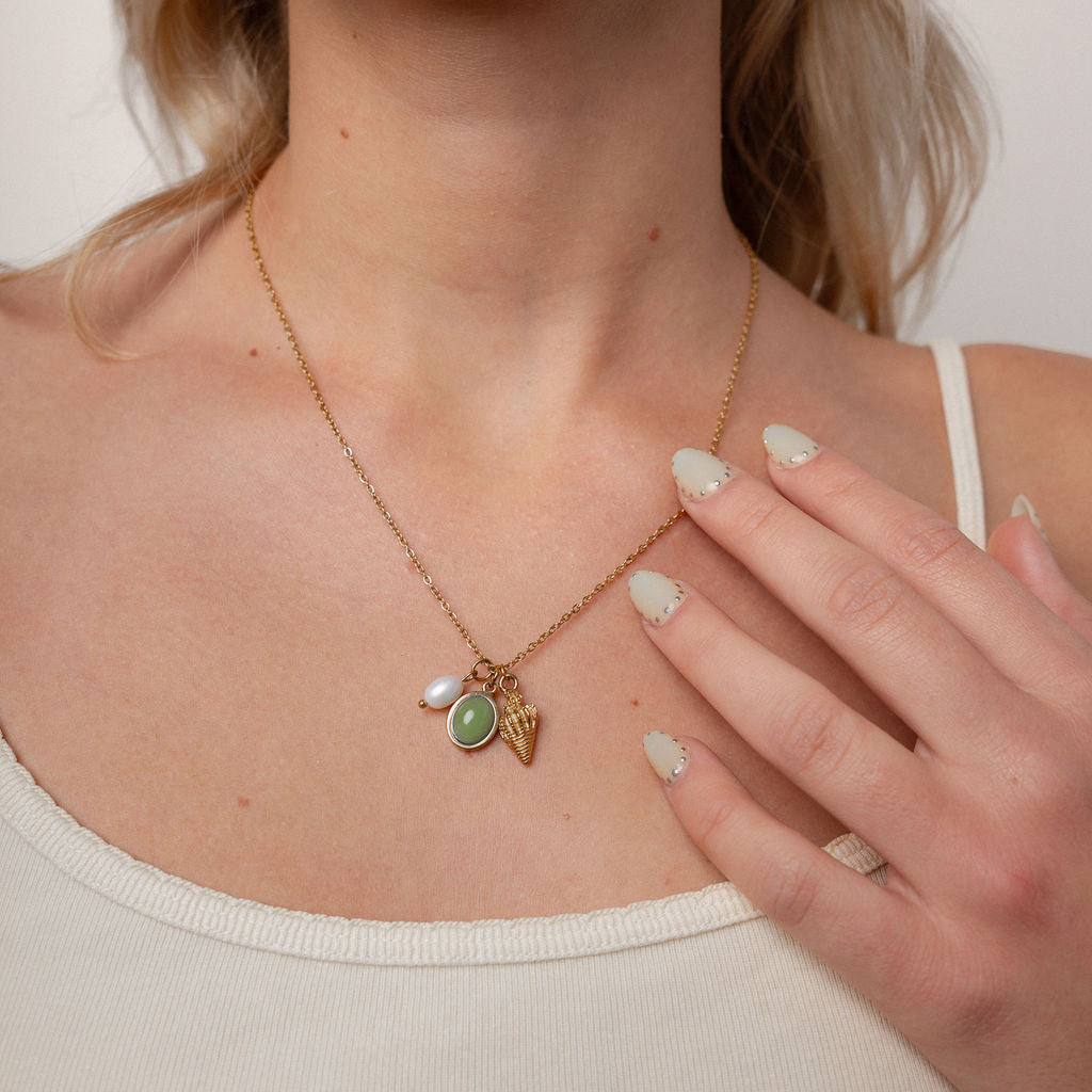 A close-up of a woman wearing a delicate gold necklace with a small pendant featuring a green oval stone and a white pearl, paired with a gold ring.