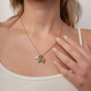 A close-up of a woman wearing a delicate gold necklace with a small pendant featuring a green oval stone and a white pearl, paired with a gold ring.