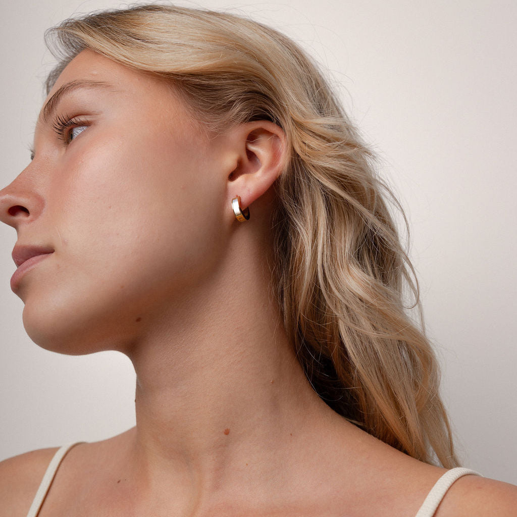 A close-up side profile of a blonde woman wearing a small gold hoop earring against a plain light beige background.