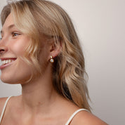 A close-up side profile of a smiling blonde woman wearing a small gold hoop earring with a white pearl drop, paired with a cream-colored spaghetti strap top against a plain light gray background.