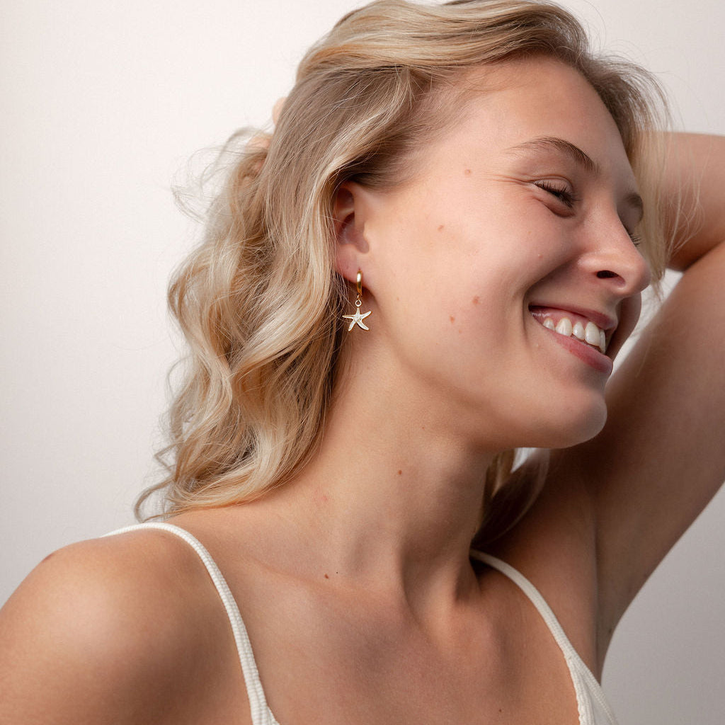 A smiling blonde woman wearing a delicate gold star-shaped stud earring, shown in a close-up portrait against a plain light background.