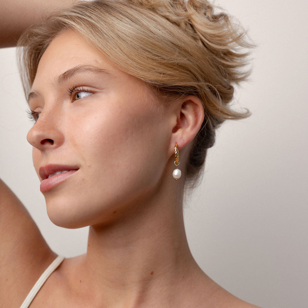 A close-up of a woman wearing a delicate gold dangle earring with a white pearl drop, shown in profile against a plain light background.