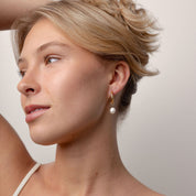 A close-up of a woman wearing a delicate gold dangle earring with a white pearl drop, shown in profile against a plain light background.