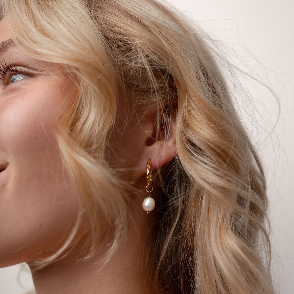 A close-up side profile of a woman with blonde hair wearing a delicate gold dangle earring with a single white pearl drop, set against a plain light beige background.