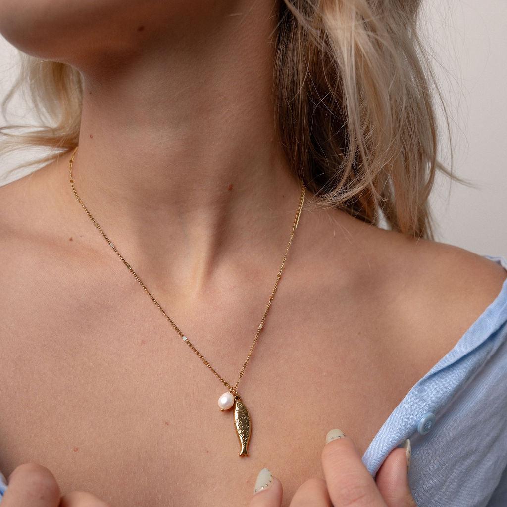 A close-up of a woman wearing a delicate gold necklace with a small leaf-shaped charm and a pearl accent, paired with a light blue off-the-shoulder top.