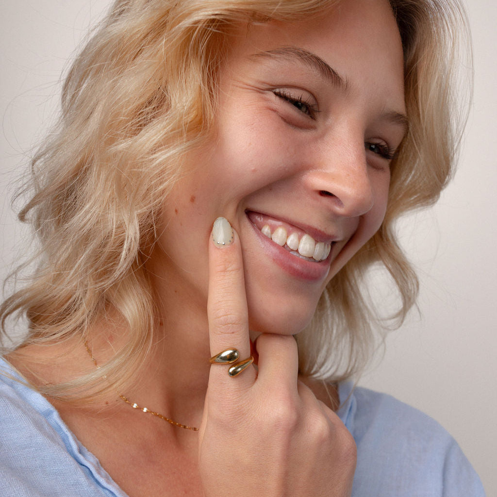 A close-up lifestyle shot of a smiling blonde woman wearing a gold snake ring on her finger, a delicate gold necklace, and a light blue top against a plain light background.