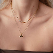 A close-up of a model wearing a double-layered gold necklace. The shorter necklace features a polished gold whale tail pendant, while the longer necklace has small gold beads with white pearl accents.