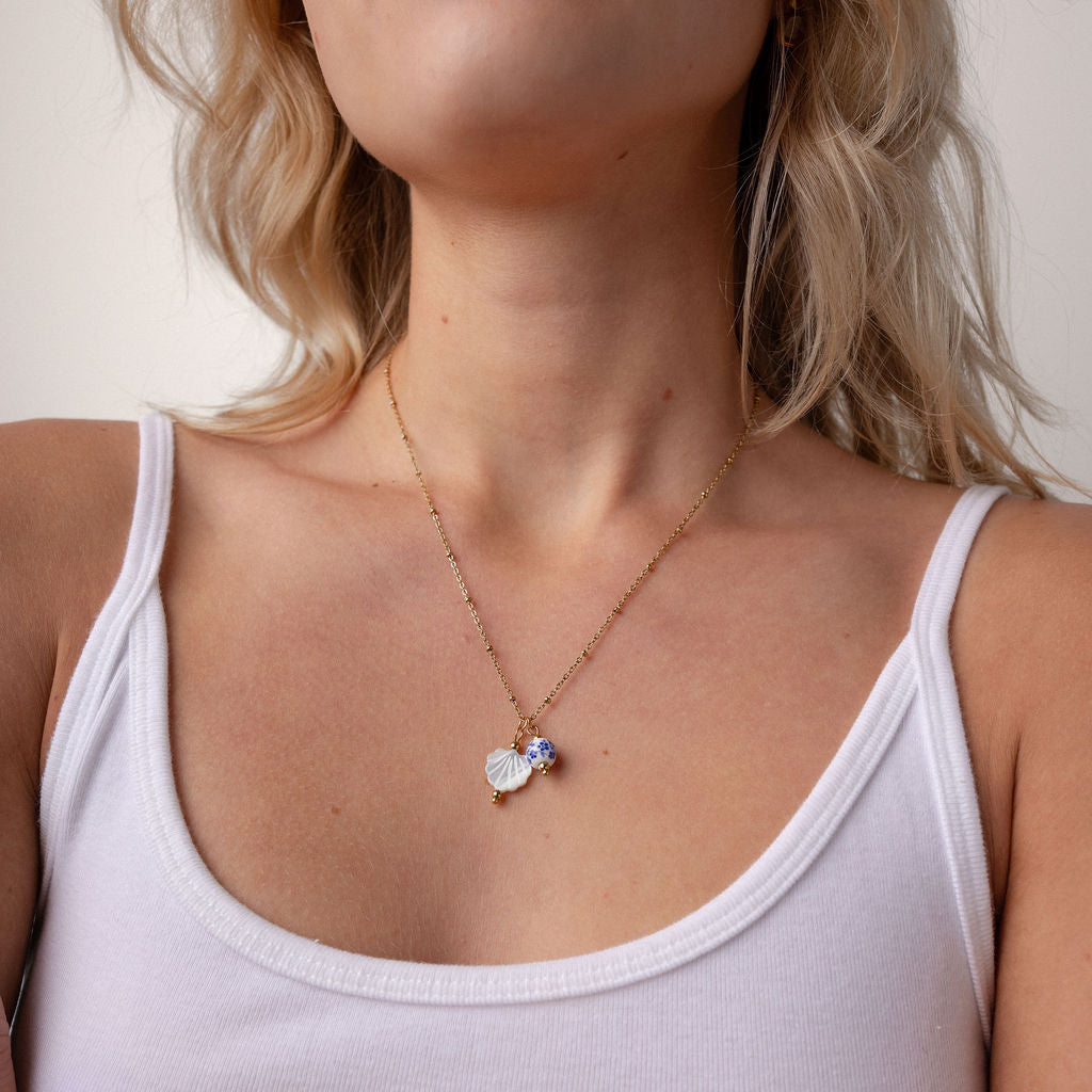 A close-up of a woman wearing a delicate gold chain necklace with a small heart-shaped pendant and a tiny blue gemstone accent, styled with a white ribbed tank top.