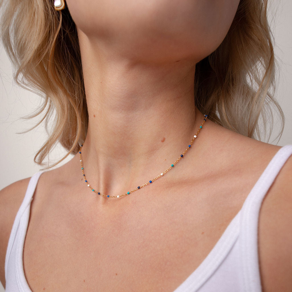 A close-up of a woman wearing a delicate multi-colored beaded necklace with a small gold-toned pendant, paired with a white tank top.