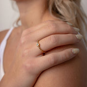 A close-up lifestyle shot of a woman wearing a delicate gold ring with a small clear gemstone on her finger, with a matching gold drop earring visible. The background is a soft, neutral beige.
