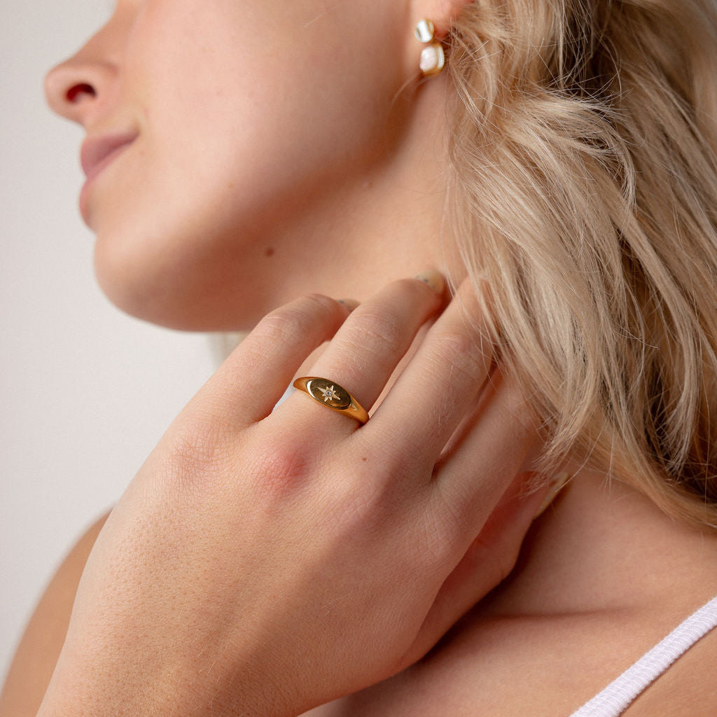 A close-up lifestyle shot of a woman wearing a gold signet ring on her finger and a small gold hoop earring in her ear, set against a soft beige background.