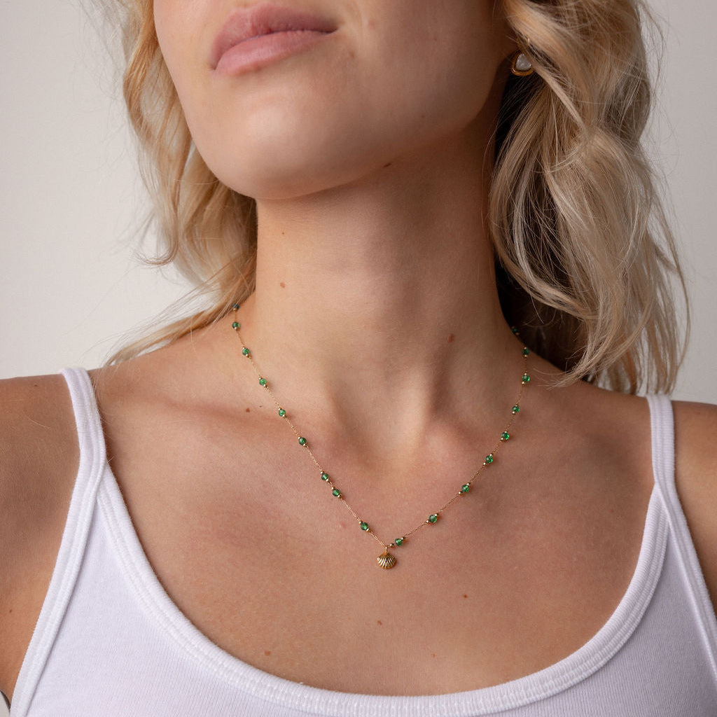 A close-up of a woman wearing a delicate gold necklace with small green bead accents and a tiny gold charm, paired with a white ribbed tank top.