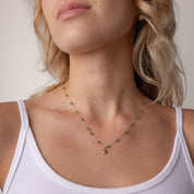 A close-up of a woman wearing a delicate gold necklace with small green bead accents and a tiny gold charm, paired with a white ribbed tank top.