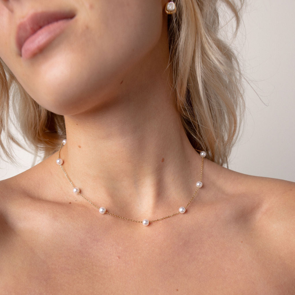A close-up of a model wearing a delicate rose gold necklace with small white freshwater pearls spaced along the chain, paired with a dark brown strapless top.