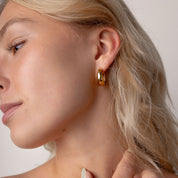 Close-up of a woman wearing a small gold hoop earring and a thin gold ring on her finger, shown in profile against a soft light beige background.