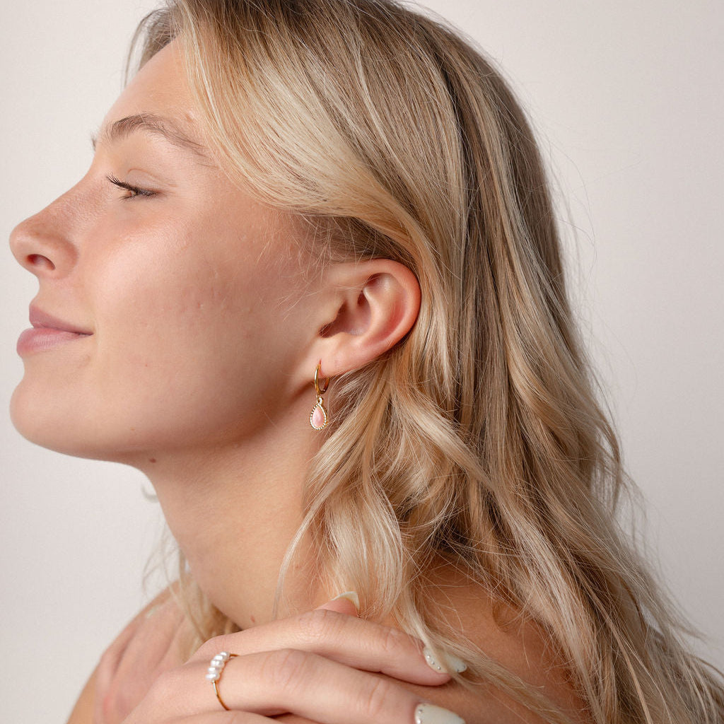 A close-up side profile of a woman with blonde hair wearing delicate gold jewelry, including a small hoop earring and a ring on her finger, against a plain light background.