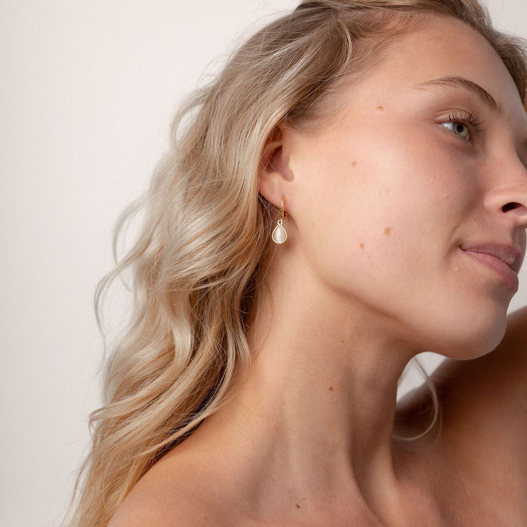 A close-up side profile of a blonde woman wearing a small, delicate gold hoop earring with a white pearl drop, set against a plain off-white background.