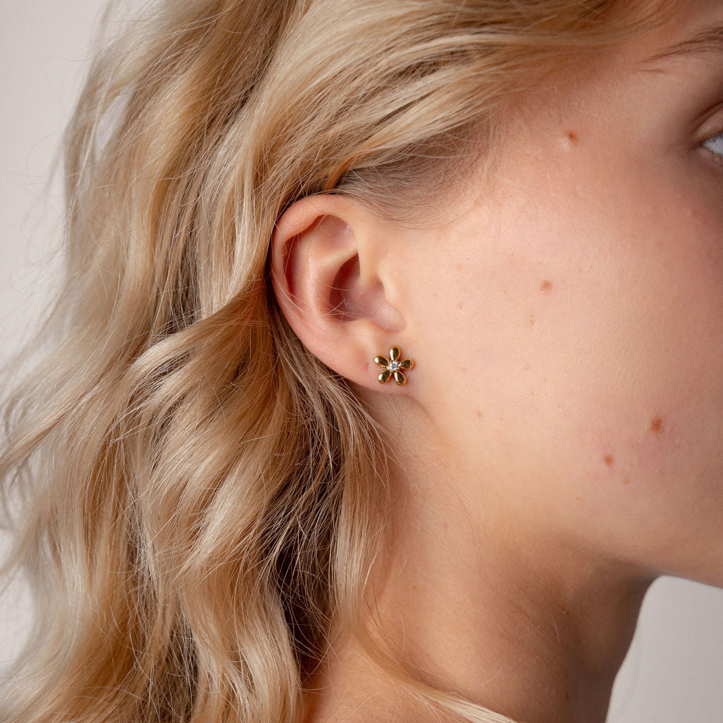A close-up side profile of a woman with blonde hair wearing a small gold flower-shaped stud earring.