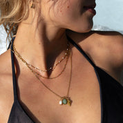 A close-up lifestyle shot of a model wearing a layered gold necklace with a green and white shell pendant over a black bikini top in water.