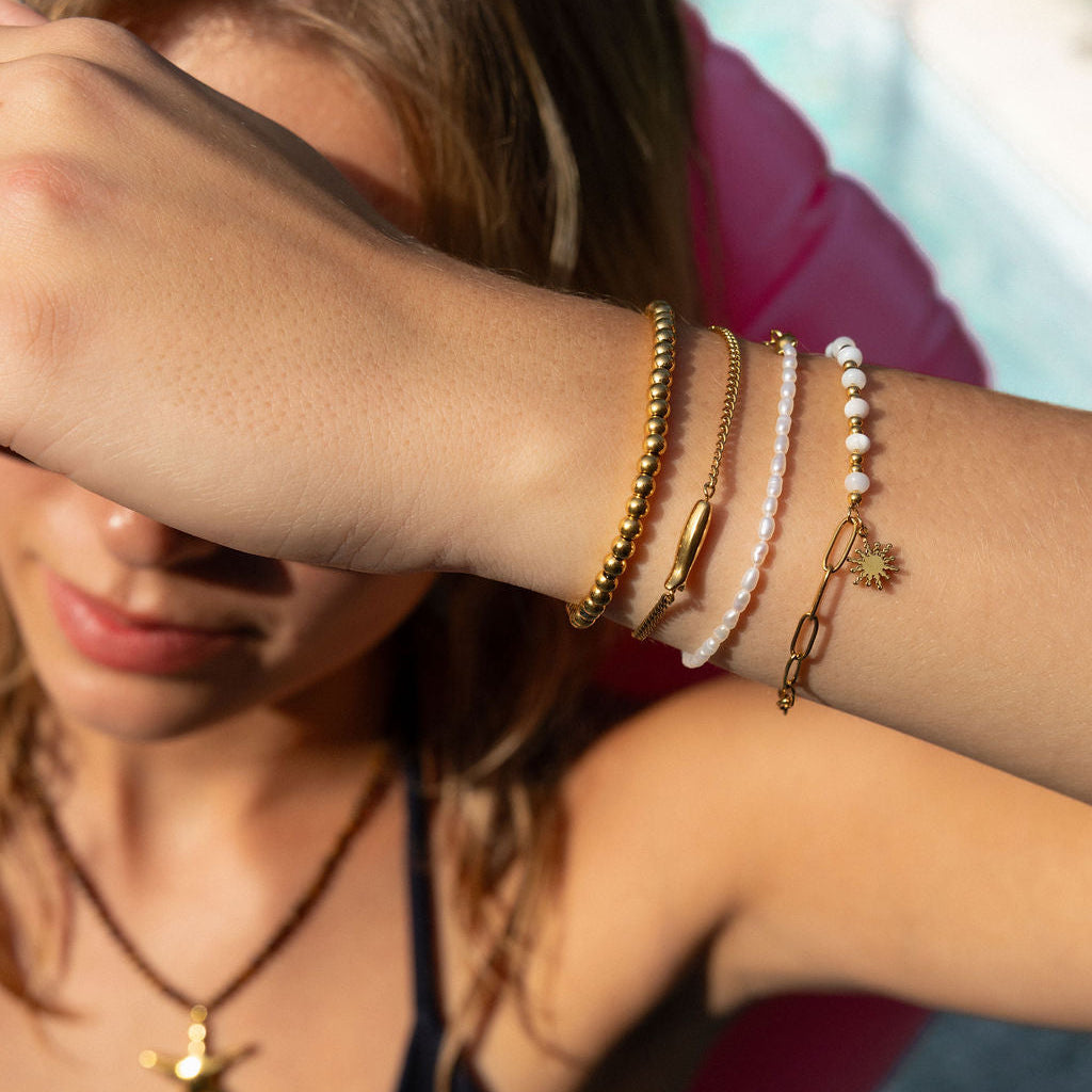 A close-up lifestyle shot of a woman's wrist adorned with multiple delicate bracelets, including gold chain, pearl bead, and rope bracelets, while she wears a black swimsuit and a starfish necklace.