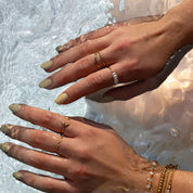 A close-up of two hands and wrists wearing multiple delicate gold rings and bracelets, partially submerged in rippling water. The jewelry includes thin gold bands, a small star-shaped ring, a pearl-studded ring, and stacked beaded bracelets.