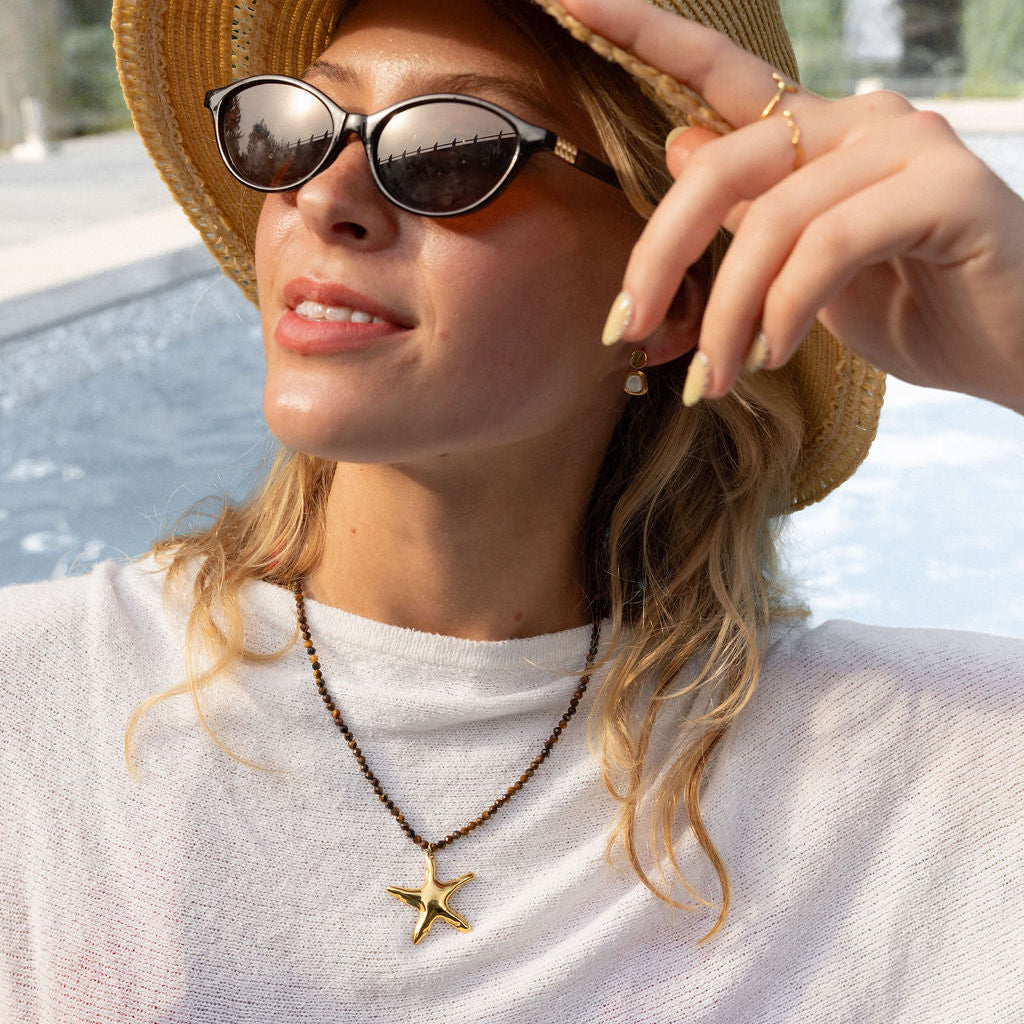 A woman wearing a straw sun hat, dark sunglasses, and a gold starfish pendant necklace, posing outdoors near a pool.