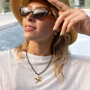 A woman wearing a straw sun hat, dark sunglasses, and a gold starfish pendant necklace, posing outdoors near a pool.