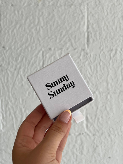 A hand holding a white jewelry box with the text 'Sunny Sunday' on it.